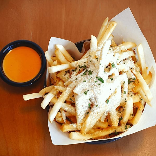 A basket of golden french fries topped with parmesan cheese and parsley, with a side of orange dipping sauce.