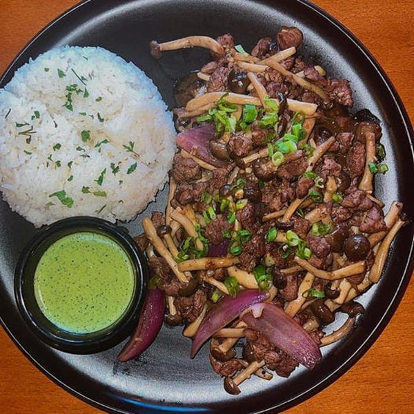Vietnamese beef and mushroom stir-fry with rice and green dipping sauce on a black plate.